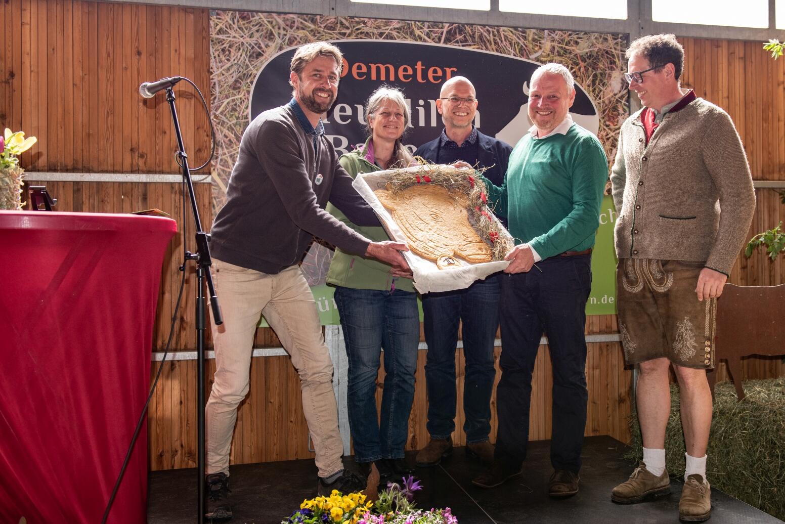 Christoph Reiber (Geschäftsführer Demeter Baden-Württemberg), Ute Haimerl (Geschäftsführerin Demeter Bayern) und Alexander Gerber (Vorstand Demeter e.V.) überreichen Rolf Holzapfel und Andreas Aufmuth (beide Erzeuger bei den Demeter HeuMilch Bauern) auf der Jubiläumsfeier in Ankenreute einen großen Keks in Form einer Kuh – gebacken von Demeter-Bäckermeisterin Anne Dorer von der Eselsmühle.